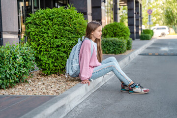 A beautiful teenage girl with a backpack is sitting on the road  before school. The concept of...