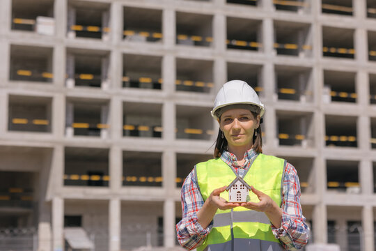 woman engineer in a white helmet and a protective vest against the background of a house under construction holds a miniature model of the house in her hands. construction sale and purchase of real
