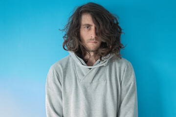 Serious long haired young man in grey hoodie on blue background