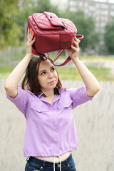 Fototapeta na wymiar Young woman on street in rain. Positive female in purple shirt holding backpack over head, sheltering from rainfall.