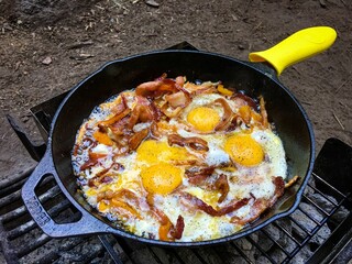 Delicious breakfast during camping with eggs and bacon sizzling in an iron pan over charcoal