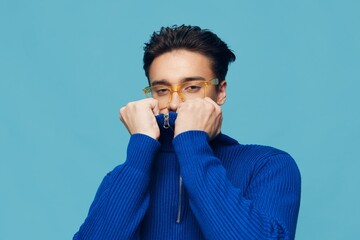 a cute, handsome man stands on a light blue background in a blue zip-up sweater with yellow sunglasses on his face and looks at the camera, turning up his collar and hiding his face