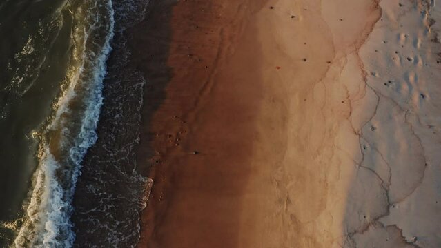 Bird's-eye view of a sandy deserted beach washed by foamy sea waves. View from above, turning into a panoramic shot of the coastline.