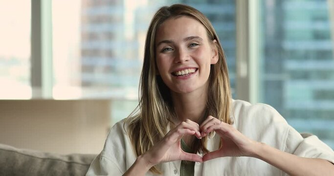Positive beautiful adult girl showing finger heart shape from joined hands at camera, getting happy, smiling, laughing. Young blogger woman sharing support, gratitude, love, making like sign