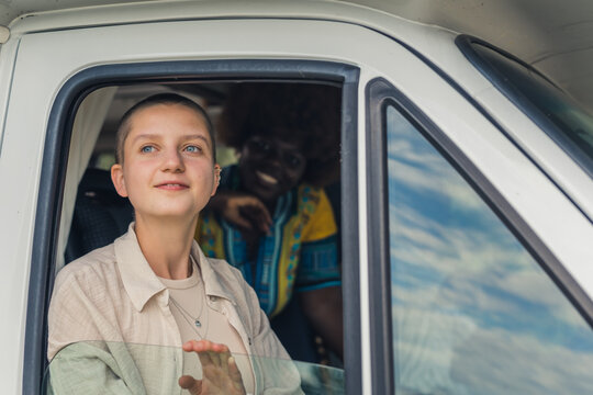 Young caucasian beautiful girl with shaved head looking out of the open window of a camping van in awe. Her smiling African young woman friend with kinky hair on the back seat. High quality photo