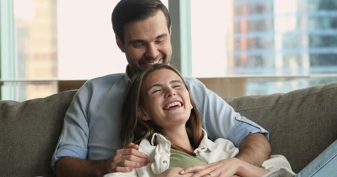 Happy young dating couple in love enjoying leisure at home, relaxing together on couch, looking away, thinking, dreaming, talking, laughing, discussing plans for family. Guy hugging girlfriend on sofa