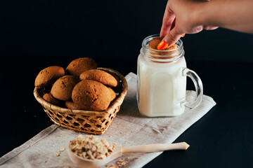 Basket of fresh oatmeal cookies with milk in jar, raw oat flakes in large wooden spoon on napkin on black background. Unrecognizable woman dips cookie in milk.