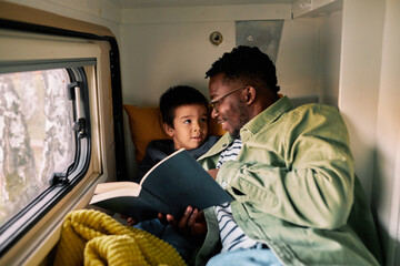 Multicultural father reads a book to his son in a van. - 526390416