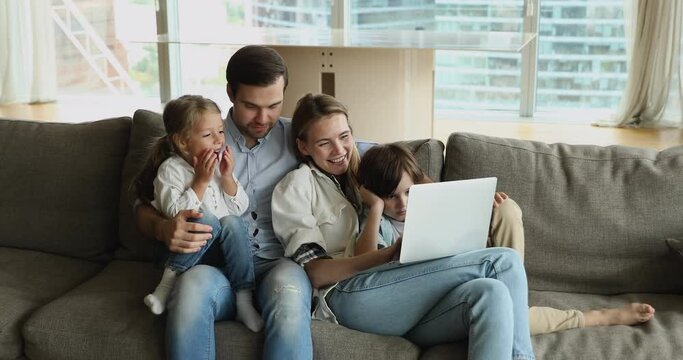 Happy family using laptop together on comfortable couch. Couple of cheerful parents and two cute kids using online learning app in computer, hugging, talking, laughing, enjoying leisure