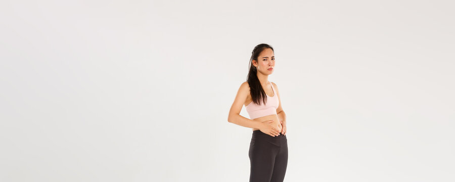 Full length of upset and gloomy asian fitness girl trying to lose weight, touching belly, cant see results from workout or diet, feeling uneasy while standing in sportswear over white background