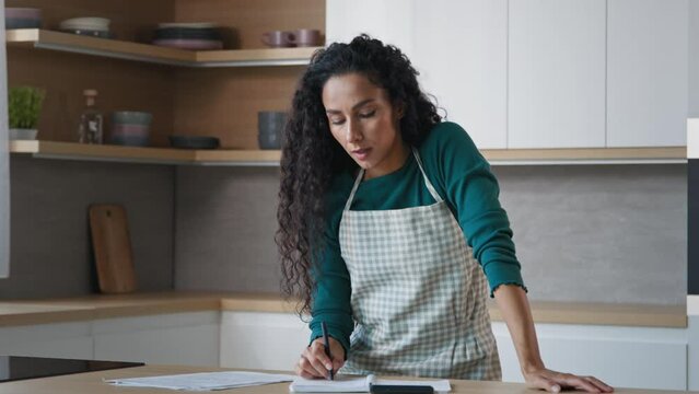 Arabian hispanic housewife pretty curly woman wears apron stand in kitchen write notes food recipe use calculator for counting calories checklist of products calculate domestic bills paperwork at home