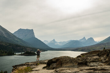 Man Stands On Cliff Edge ane Photographs Saint Mary Lake