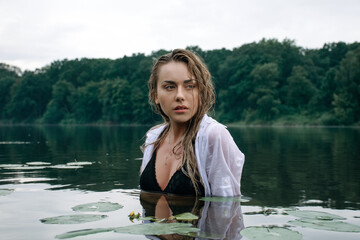 Beautiful sensual blonde girl with wet hair in a white shirt and black swimsuit posing in the lake after the rain.