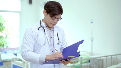 Asian doctor reading a medical document on notepad in hospital treatment ward. Health care and wellbeing concept.