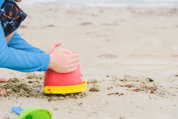 Child building a sand castle on the beach with a red bucket on a cloudy day