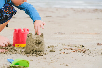 Child building a sand castle on the beach with a red bucket on a cloudy day