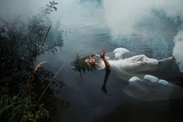 A lake nymph in a white dress and a wreath of flowers floats on the surface of the water. A young woman in a folk image, a mystical story - 526410879