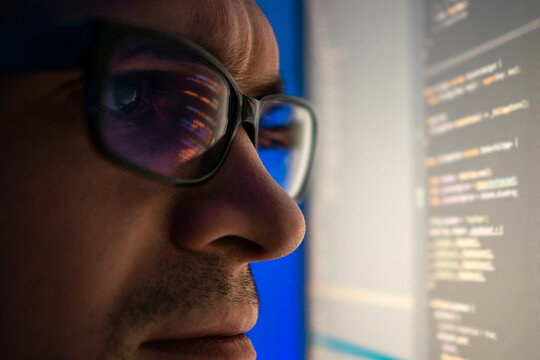 A programmer, a man in front of a computer, the code is reflected in the glasses. Working in IT, learning programming languages, developing applications and software.