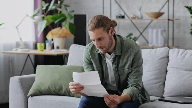 Unquiet young caucasian man opening paper envelope, reading letter with bad news. Angry handsome freelancer received notification about high bills, dismissal notice, bank debt, tax invoice.