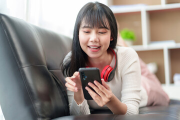 Pretty and cute Asian woman relaxing on the sofa, playing social media and accessing apps various entertainments, wear headphones, listen to music happily at home.