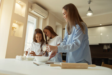 Fototapeta premium Happy family preparing food together in the kitchen. Mom teaches her daughters how to cook and knead the dough. Mother's Day concept. pour milk