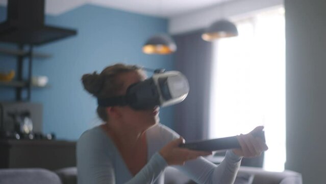 Young futuristic blonde girl wearing virtual reality headset, holding controller playing shooter video game at home. Woman playing avatar VR shooting fight in metaverse in the middle of a living room.