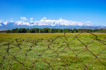 Fototapeta na wymiar A metal lattice fence is installed on a plain against the backdrop of mountain peaks covered with snow. A beautiful landscape is made by a tourist while traveling in the mountains. 