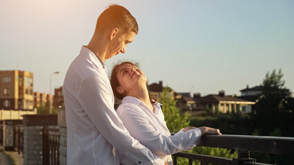 Young couple with cerebral palsy wearing white shirts looks at each other smiling and hugging. Man and woman enjoy resting together against blurry buildings