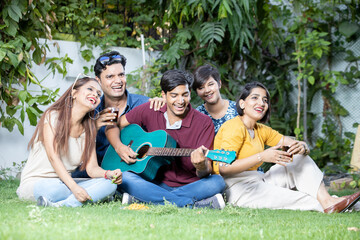 Happy young indian friends sitting at park or garden playing guitar and music having fun together and enjoy outdoor picnic.