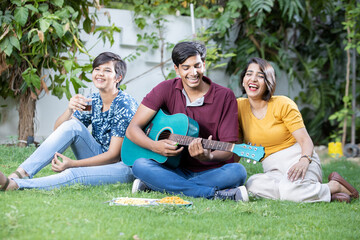 Naklejka premium Happy young indian friends sitting at park or garden playing guitar and music having fun together and enjoy outdoor picnic.
