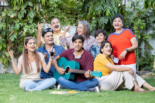 Portrait Of Happy Multi Generational Indian Family Sitting Enjoy Picnic Together Outdoor Garden Playing Guitar And Music Having Fun.