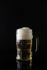 Light beer with foam in a transparent glass on a mirrored black table. Black background. Golden beer in a faceted mug
