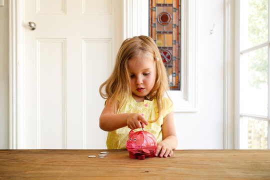 Blonde girl wearing yellow blouse putting a coin in a pink transparent piggy bank