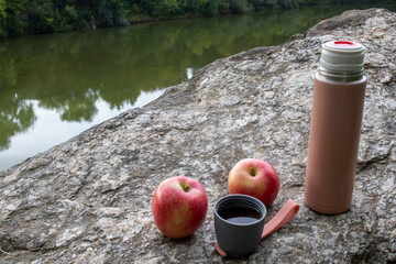 Beige thermos bottle, cup of tea or coffee, and red apple on rock over the river. Beautiful summer, spring, autumn landscape. Picnic on the rock - obrazy, fototapety, plakaty