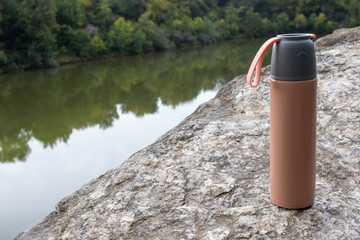 Beige thermos bottle and cup of tea or coffee on rock over the river. Beautiful summer, spring, autumn landscape. Picnic on the rock - obrazy, fototapety, plakaty