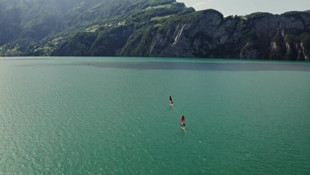 Two men are windsurfing on a picturesque lake at the foot of the Alps in Switzerland. Drone point of view, panoramic shot from the side of the lake