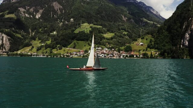 Scenic drone footage of a sailing boat with a Swiss flag sailing on a beautiful lake at the foot of the Alps, not far from a village located on the coast
