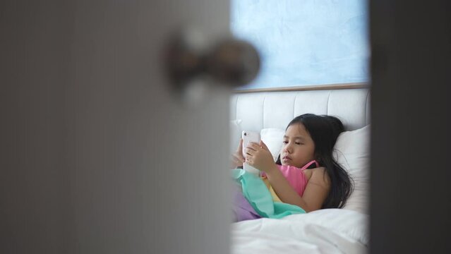 asian child or kid girl sleep lying to pillow on bed for playing looking smartphone or addicted mobile phone to hyperactive with parents secretly watch to daughter or baby behind ajar door in bedroom