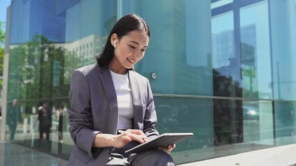 Happy young Asian business woman executive, female professional wearing suit holding digital tablet using software applications working online sitting in big city near glass business center outdoor.