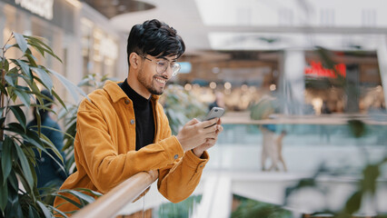 Young hispanic guy stands in mall holding phone smiling sending email on social network surfing web...
