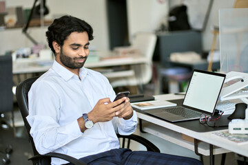 Indian young business man company worker, professional employee, stock market trader holding smartphone using cell phone mobile apps reading news, checking financial market data at work in office.