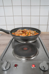 Electric stove in the kitchen with a frying pan cooking chicken legs and vegetables, with copy space background. Concept food and economic crisis