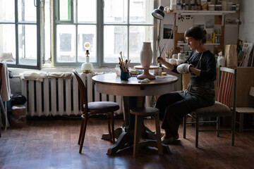Female craftsperson in apron enjoys working with pottery craft in workshop. Young woman sits on chair at round table with equipment against bright window painting handmade ceramic vase with brush