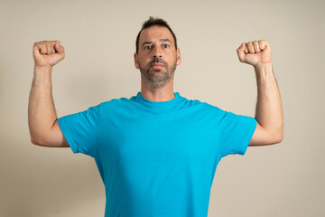 Fototapeta premium Caucasian happy unshaven strong sporty fitness student male 40s in casual blue t-shirt showing biceps muscles on hand isolated on beige background studio portrait