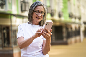 Mature woman with modern technologies. Senior grey hair lady holding smartphone standing outdoor at the streets of old town. Mature woman read text message standing or walking outdoors