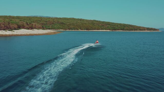 Young man in red swimming suit enjoying his holiday riding electric surfboard in a calm blue ocean bay between yachts and sailboards. Summer sport for friends.