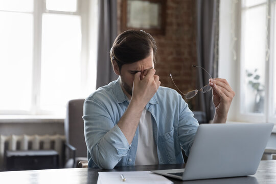 Tired millennial businessman sitting at laptop, rubbing eye lids, taking glasses off, touching head, feeling exhausted, overworked, suffering from weak eyesight, headache, high blood pressure