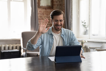 Happy millennial freelance business man in casual talking to client on video call, waving greeting hand, saying hello, using tablet computer at work table, getting good news, smiling, laughing