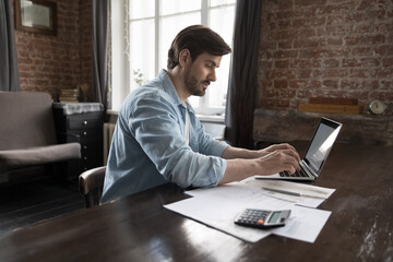 Focused freelance business man doing accounting job at home office workplace, using laptop, typing...