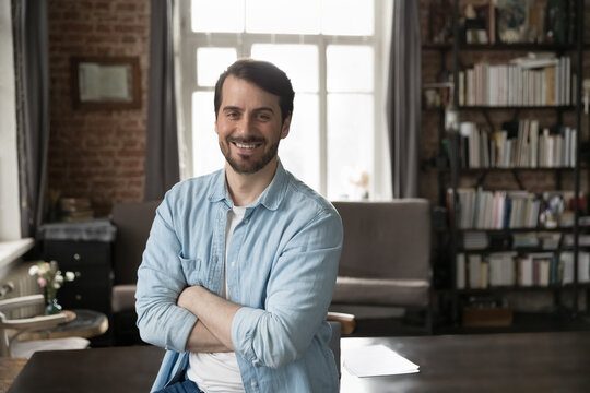 Happy confident millennial businessman in casual standing with arms crossed at work table in home office, looking at camera, smiling. Male freelance professional, business owner head shot portrait